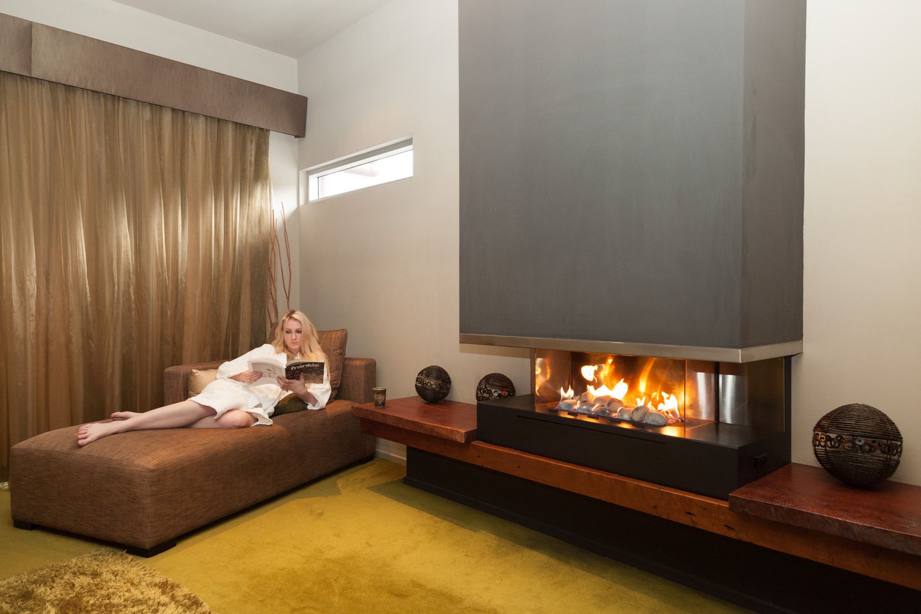 Woman in a bathrobe reading on a chaise beside a gas fireplace