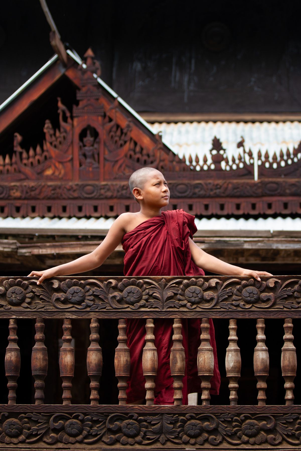 Young monk in maroon robes leaning on a carved teak balcony
