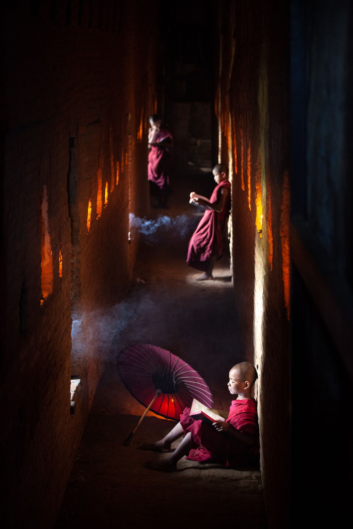 Young monks reading in a dark temple corridor lit by candles and smoke, a red umbrella beside them
