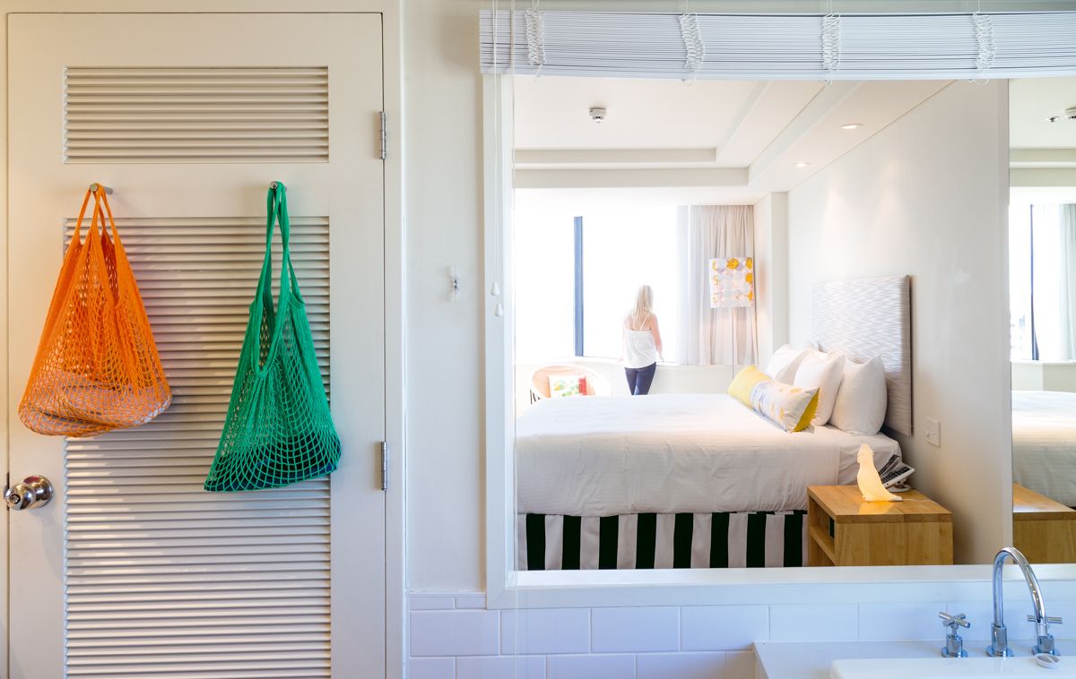 View through a bathroom window into a bright hotel room with a guest at the window