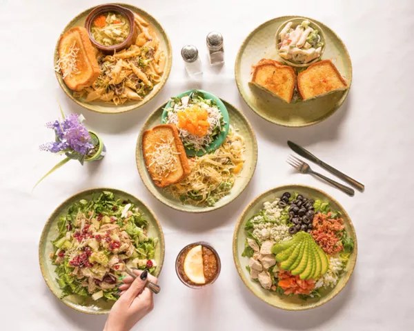Overhead view of five plates of pasta and salads on a white cloth, a hand reaching in